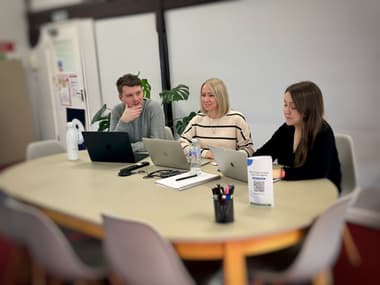 Meeting to discuss website design and development with three people: two woman and one man, all with laptops sitting at a boardroom table. On the table are two water bottles, a notepad, pen, cables, pot of pens and a standing advert with a QR code on it. In the background is a plant and there are beams on the wall. There is a door on the far top left, exiting out of the office.