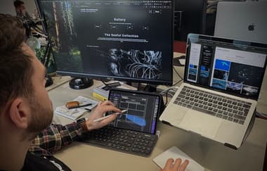 A person with short brown hair and a beard is sitting at a desk, focused on a digital tablet while using a stylus. The tablet displays a design interface, with blue elements and a keyboard below it. Two computer screens are visible: the left screen shows a dark-themed website for a nature photographer named Gabe Hearnshaw, featuring a gallery section and images of water and landscapes. The right screen displays a collection of photographs, with images of birds and water scenes.