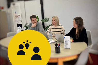 Meeting to discuss ideas between three team members at a table with open laptops and plants in the background. On the bottom left is a large yellow circle containing two black silhouette icons and a black lit lightbulb next to one of them.