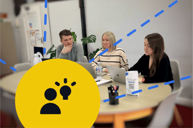 Three people are seated around a circular table in a bright, modern office space. A man with short brown hair, wearing a grey sweater, is thoughtfully resting his chin on his hand. Next to him, a woman with long blonde hair, dressed in a striped top, is looking at the table. A woman with long dark hair, wearing a black top, is focused on her laptop. In the foreground, a large yellow circle features an icon of two people and a light bulb, symbolising ideas
