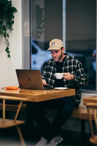 A man sits at a wooden table in a café, working on a laptop while holding a cup of coffee. He wears a checked shirt over a black t-shirt, paired with jeans and a beige cap. A small orange object is placed on the table next to the laptop. Green plants are visible in the background, along with a window showing a blurred street scene outside.