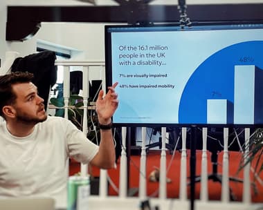 A man is talking and pointing at a screen with text on it and a graph displaying 'Of the 16.1 million people in the UK with a disability... 7% are visually impaired, 48% have impaired mobility'