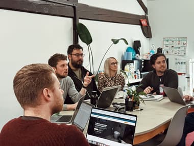 The team at WCD sits round a table, with Dean pointing at the screen where everyone else is looking. Team members are engrossed, some with laptops out in front of them.