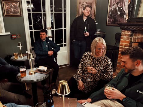 A group of five people sits in a cosy bar setting with a green wall and framed black-and-white photographs. In the foreground, a woman in a leopard print top smiles at a man in a green jacket, who holds a drink. Nearby, a man in a dark blue puffer jacket sits with his arms crossed, looking contemplative. In the background, a man with a beard stands by a door, holding a bottle. Two small tables with gold lamps are visible, one with a pint of beer on it.