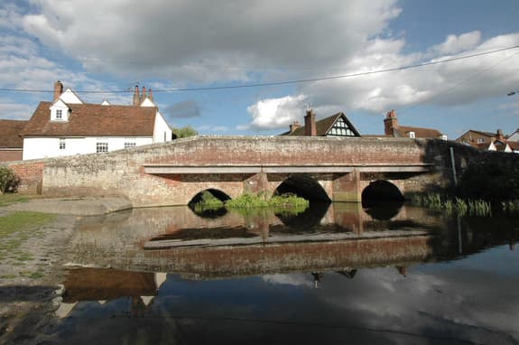 Bridge in Coggeshall, with the river passing through it and houses seen from behind. It is a blue and cloudy day.
