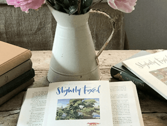 A pitcher with flowers on a wooden table with a book in front of it opened up, and a copy of Slightly Foxed on top.