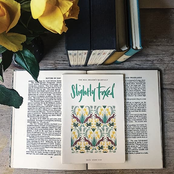 A wooden table with an open book and a copy of Slightly Foxed on top. Behind are some more books standing upright, and yellow roses are in the top left of the image.