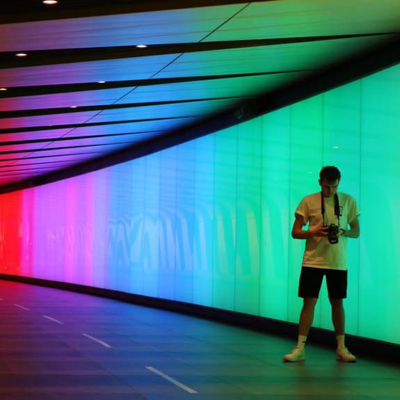 A man stands inside a tunnel that has panels running along it with different lights, showing rainbow colours. He is wearing a light t shirt, shorts, socks and trainers. He has a camera aroudn his neck and is looking at the camera screen.