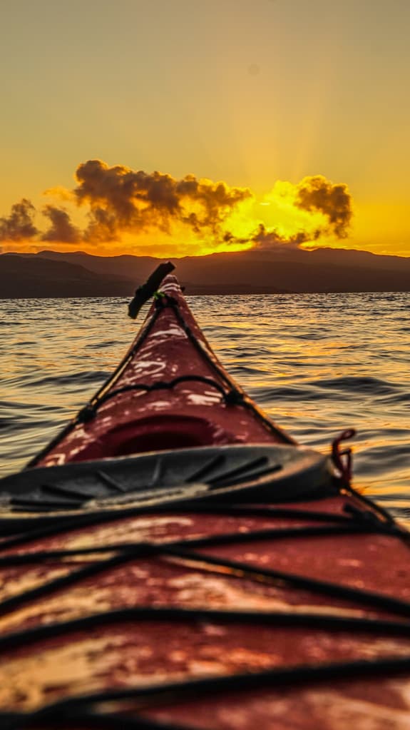 A photo is taking from the person sitting inside a red kayak, looking out from the sea over to mountains, with the sun setting behind them