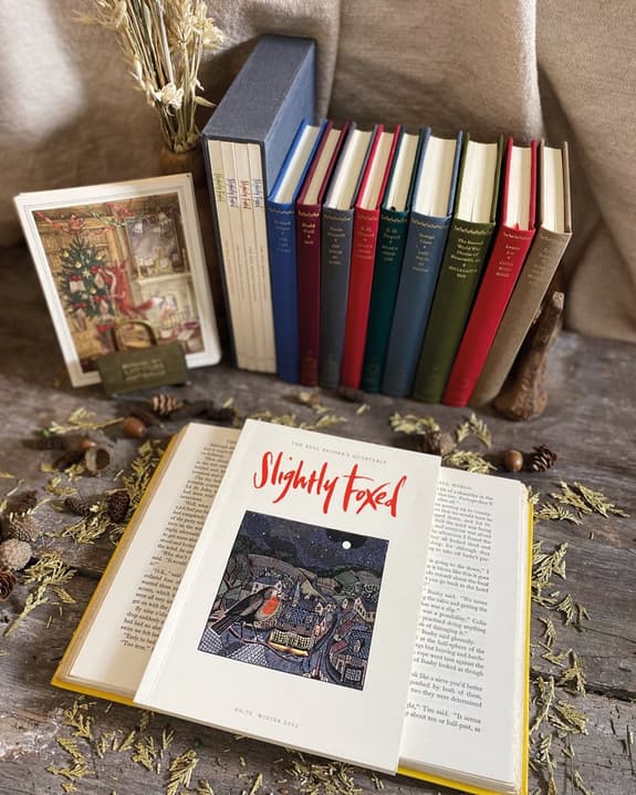 The image features two books on a wooden surface, one is open and one is shut. In the background are more books standing upright. Around the books on the wooden surface are dried botanicals