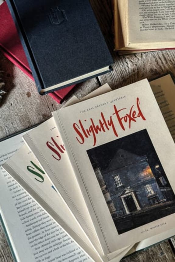 The image features a collection of books laid out on a wooden surface. Prominent in the foreground is a book titled "Slightly Foxed," which has a cream cover with red lettering and a dark, illustrated image of a house. Surrounding this book are several other books, including one with a black cover and a red spine, partially visible at the top left. Other pages are open, displaying text in a light cream colour. The background includes more books, creating a warm and inviting reading atmosphere