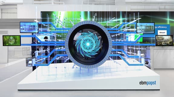 A modern exhibition display featuring a large circular structure at its centre, designed to resemble a turbine or fan, with a swirling blue and white graphic at its core. Surrounding the circular element are transparent panels and blue lines extending outward, creating a dynamic visual effect. In the background, several screens display images and information related to energy efficiency. Two people in business attire are visible near the display, engaging with the content. The logo "ebmpapst" is prominently featured on the front of the display