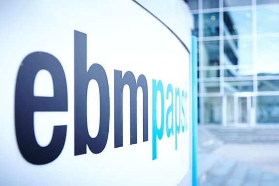 A close-up view of a sign displaying the word "ebmpapst" in a modern font. The letters "ebm" are in bold black, while "papst" features a lighter blue colour for the letter "p" and remains in black for the remaining letters. The background shows a blurred, contemporary building with glass windows and steps leading up to the entrance. The overall atmosphere is professional and clean, suggesting a corporate environment.