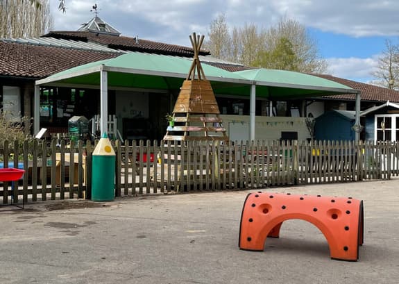 A school playground with various equipment shown including a wooden teepee and a bin in the shape of a pencil