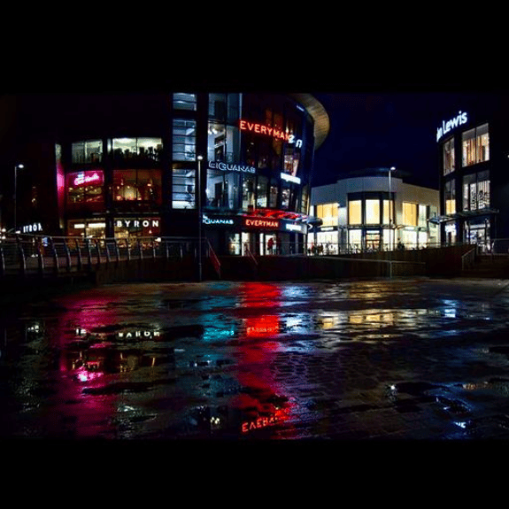 A night-time urban scene in Chelmsford featuring a modern building with large glass windows. The building displays illuminated signs for "Everyman" and "Byron," while a nearby structure features "John Lewis." The ground is wet, reflecting vibrant colours from the lights, including reds, blues, and yellows. The scene captures a lively atmosphere, with people visible in the distance, and the overall setting suggests a bustling shopping area.