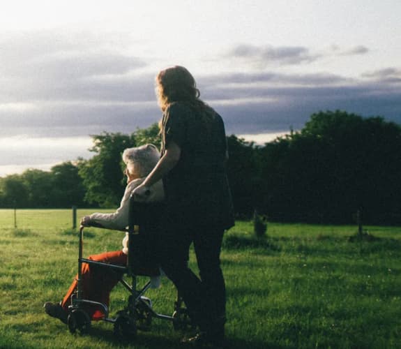 A woman with mid length hair is in a field pushing along a wheelchair with an older woman with grey hair