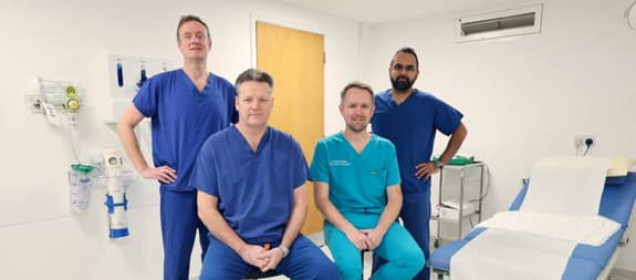 Four male doctors are in a consulting room. Two are standing with hands on hips, and the other two are seated with their hands clasped.