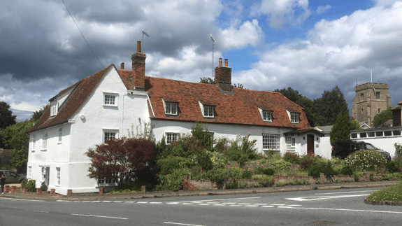 A large, white, two-storey house with a distinctive orange tiled roof is set against a cloudy sky. The house features multiple windows, and a well-kept garden with colourful flowers and shrubs surrounds its front. A road runs in front of the house, with a person standing on the pavement nearby. In the background, a stone church with a square tower is visible, surrounded by trees. The overall scene captures a quaint, rural atmosphere.