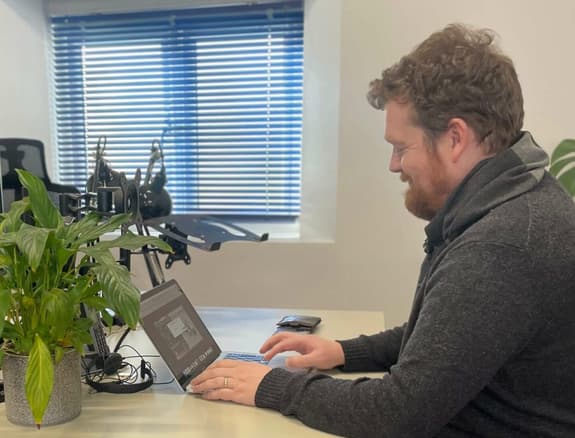 A man with red curly hair and a red beard sits at a desk with a laptop and a potted plant