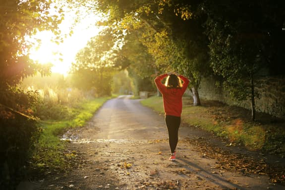 A woman walks along a country road at sunrise, with her arms on her head wearing an orange sweatshirt, orange trainers and black trousers. The sun is glowing against her outline.