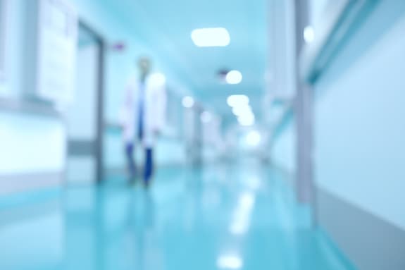A blurred hospital corridor is depicted, featuring smooth, shiny floors in a light blue hue. The walls are painted in a similar blue tone, creating a calming atmosphere. In the foreground, a person in a white lab coat and blue trousers is walking, though their features are indistinct due to the blur. Bright overhead lights illuminate the corridor, which stretches into the distance, lined with doors and windows that are not clearly visible. The overall image conveys a sense of a healthcare environment.