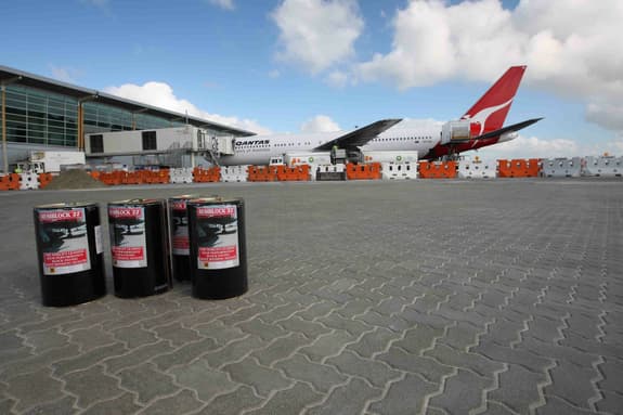 The image shows a busy airport scene with a Qantas airplane parked in the background. The aircraft is primarily white with a red tail featuring the Qantas logo. In the foreground, there are five black barrels labelled "RESIBLOCK '22," which describe a high-performance joint bonding sealer. The ground is paved with patterned grey blocks, and several orange and white safety barriers are visible behind the barrels, indicating ongoing construction or maintenance work. The sky is partly cloudy, adding to the atmosphere of the scene