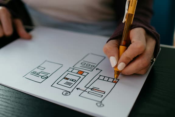 A close-up image of a hand holding a yellow pen, drawing on a sheet of white paper. The paper features a series of simple, hand-drawn diagrams illustrating a design process with numbered steps. The diagrams include outlines of boxes and rectangles, with some sections shaded in orange. The background shows a dark wooden table surface and a blurred blue object, possibly a chair or wall. The person is wearing a long-sleeved maroon top.