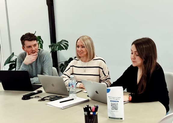 Three people are seated at a light-coloured table in a modern office space. On the left, a man with short dark hair and a grey sweater is resting his chin on his hand, looking thoughtfully at the others. In the centre, a woman with straight blonde hair, wearing a striped sweater, is smiling slightly as she engages in the conversation. On the right, a woman with long brown hair, dressed in black, is focused on her laptop. Each person has a laptop in front of them