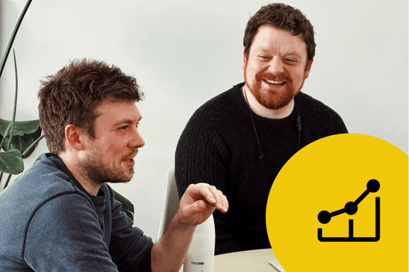 Two men are engaged in conversation at a table in a bright, minimalistic room. The man on the left has short, tousled hair and is wearing a dark blue sweater, smiling as he gestures with his hand. The man on the right has curly red hair and a beard, wearing a black sweater, also smiling. A green plant is visible in the background. In the bottom right corner, a yellow circle contains a black icon of a line graph, symbolising growth or progress.