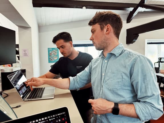 Two men are standing pointing at designs on a laptop. They're at a standing desk in a large offiec with beams on the ceiling. The brown-haired man at the front is wearing a smart watch and blue shirt, and the man at the back has darker brown hair and is wearing a navy blue t shirt with the logo 'We Create Digital' on it.