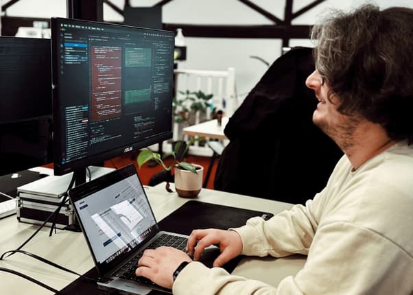 Man is smiling whilst developing a website using a laptop and computer monitor