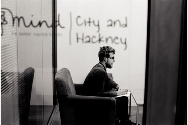 A young man with glasses sits in a grey armchair, facing a table with a notebook and pen in a softly lit room. He appears thoughtful, with his hands resting on his knees. The background features a glass wall displaying the text "Mind | City and Hackney" in a handwritten style. The overall atmosphere is calm and introspective, with a monochrome colour scheme enhancing the mood.