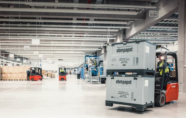 A spacious warehouse interior featuring several forklifts in operation. In the foreground, a forklift is transporting two stacked grey boxes labelled "ebmpapst" with black text, indicating ownership and weight capacity. Two workers, one in a grey shirt and another in a yellow safety vest, are near the forklifts, engaged in tasks. The background shows additional boxes on pallets, storage racks, and overhead signage, all under bright lighting. The overall atmosphere conveys a busy, organised logistics environment.