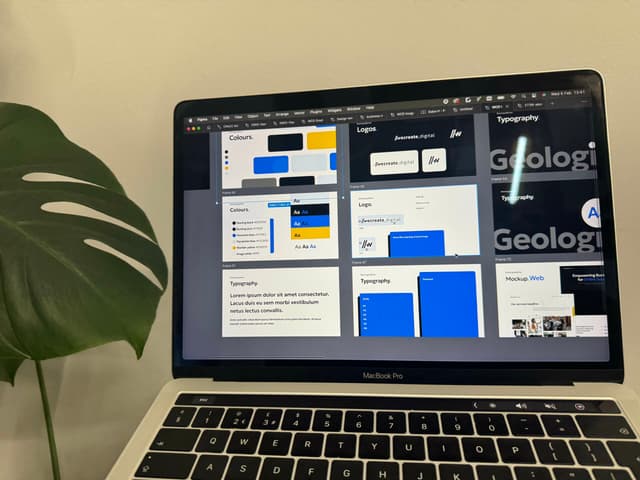 A laptop screen displays a design presentation with multiple slides. The slides feature sections labelled "Colours," "Logos," and "Typography." The "Colours" section shows a palette with various colour blocks, including shades of blue and yellow. The "Logos" slide includes examples of logo designs, featuring text and graphic elements. The "Typography" section displays different font styles, with some text highlighted. A large green monstera leaf is partially visible in the foreground, contrasting with the sleek design