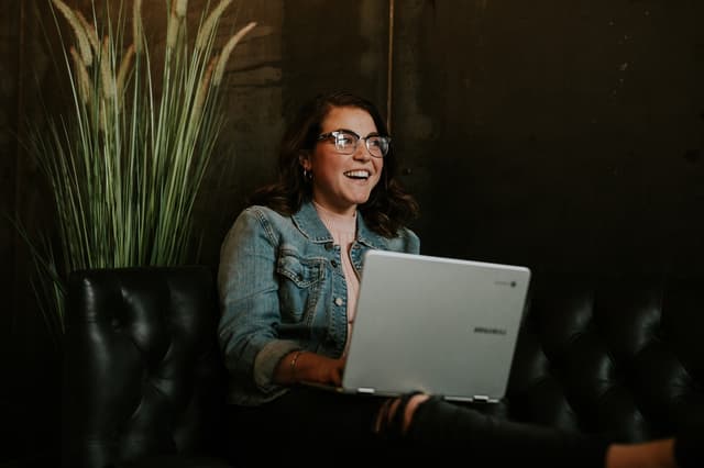 A young woman with wavy brown hair, wearing glasses and a denim jacket, sits on a black leather couch. She is smiling while using a silver laptop, which is open in front of her. In the background, there are tall green plants, adding a natural element to the setting. The wall behind her is dark, creating a contrast with her light clothing and the laptop. The atmosphere appears relaxed and inviting.