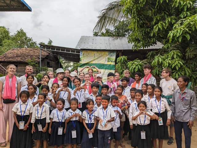 A photo set in an Asian country of lots of school children with teachers and other adults around tem. In the background are slightly dilapidated buildings and a large tree. The children are mostly smiling.