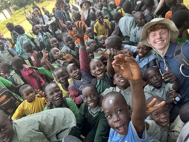 A group of children in an African country are all sitting down and smiling or laughing at the country. Other grown ups are around the edge of the children also smiling