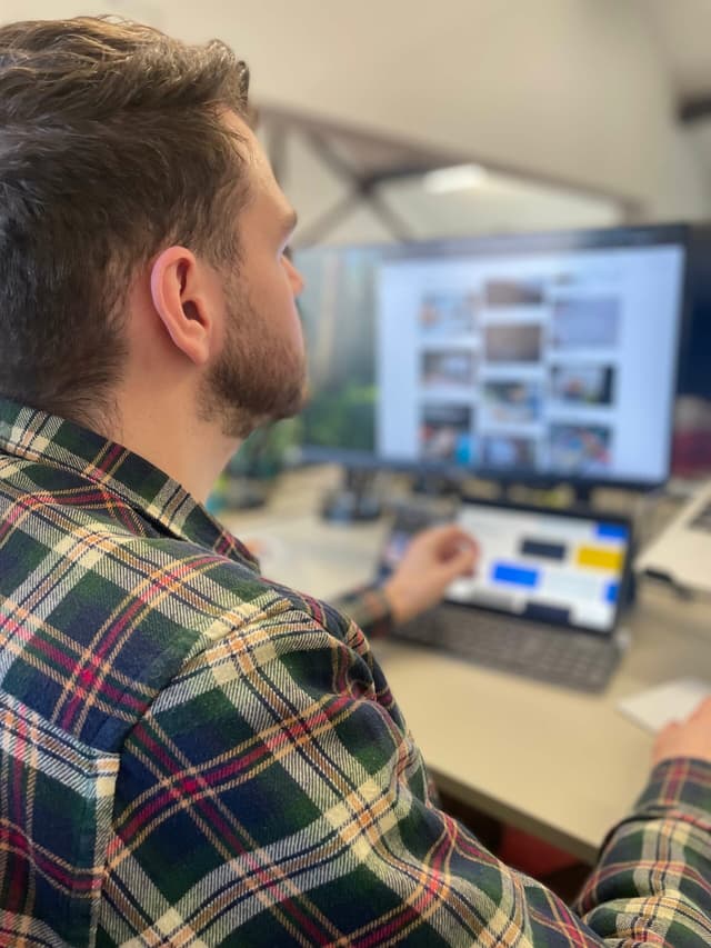 A man wearing a checked shirt is looking at a tablet and computer screen