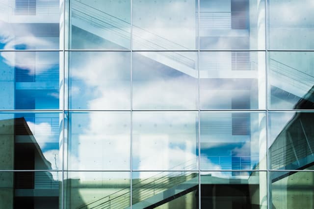 A modern glass building reflecting clouds in the sky, with visible interior staircases and railings through the transparent windows. The structure features a combination of smooth glass and concrete surfaces, creating a sleek architectural design.