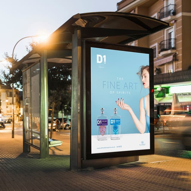 A bus stop at dusk features a large advertisement for D1 spirits. The ad background is light blue, with the text "THE FINE ART OF SPIRITS" prominently displayed. On the left, a woman with styled hair and red lipstick extends her hand, showcasing her manicured nails. To her right, two bottles of D1 spirits are illustrated: one with a blue label and the other with a decorative skull design. The bus stop is situated in an urban area, with cars visible