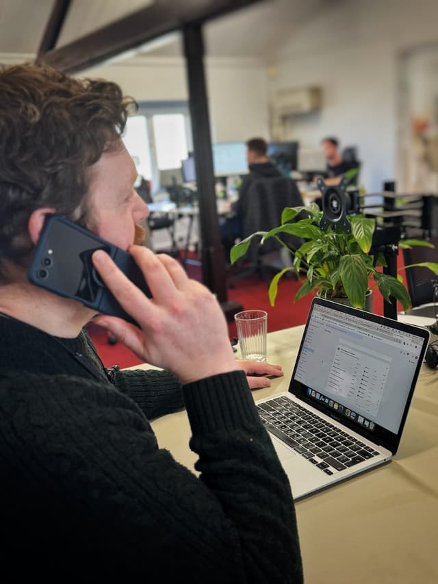 A person with curly hair is sitting at a desk, holding a smartphone to their ear while engaged in a phone call. They are wearing a dark green, knitted sweater. In front of them, a laptop is open, displaying a webpage with various data tables. A glass of water sits beside the laptop on a beige table. In the background, blurred figures of two colleagues are visible, working at their own desks with computers. A green potted plant is positioned near the laptop