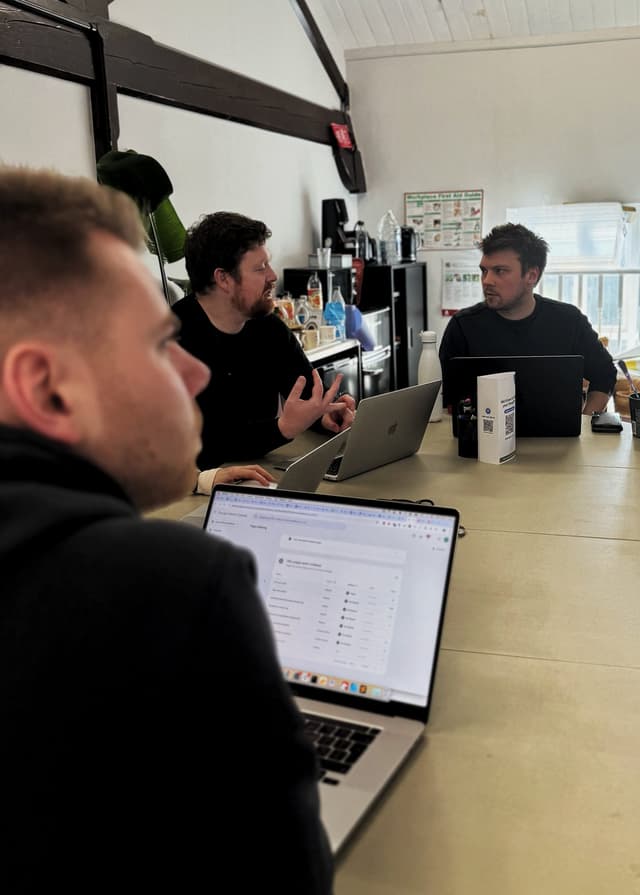 A group of three people are seated around a table in a meeting room. One person, seen from the side, is focused on a laptop displaying a spreadsheet. The second person, with a beard, is speaking and gesturing with his hands. The third person is listening intently. In the background, there are shelves with various items, including water bottles and cups. Natural light is coming from a window.