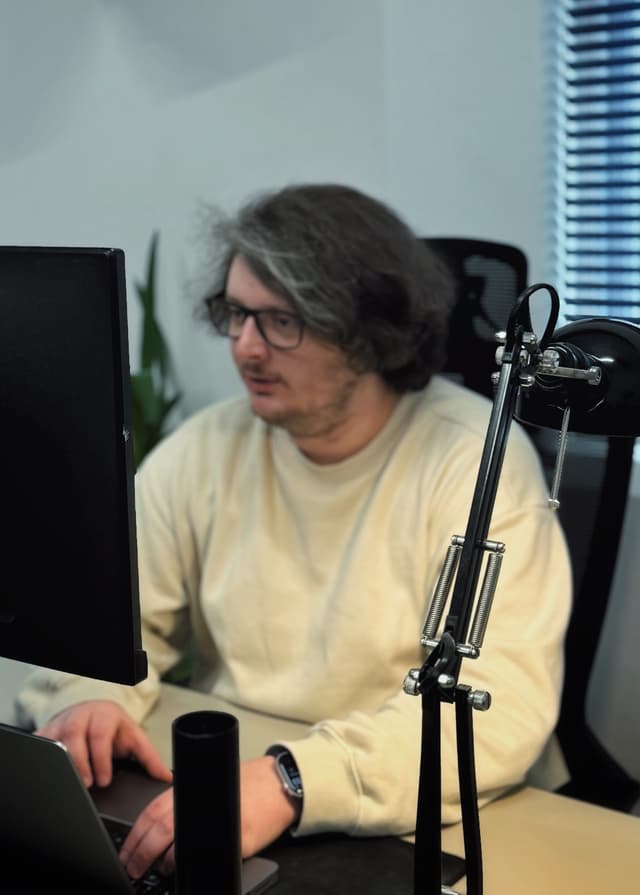 A man with wavy brown hair is sitting at a desk, focused on a laptop in front of them. They are wearing a cream-coloured sweatshirt and black glasses. To their right, there is a black desk lamp with a flexible arm, and a black cylindrical object, possibly a pen holder, is positioned on the desk. In the background, there are green plants and a window with blinds partially open, allowing light to enter the room. The overall setting appears to be a modern office space.