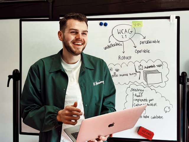 Designer standing at a white board discussing accessibility strategy. There is writing on the whiteboard mentioning WCAG 2.1 and the core principles of it.