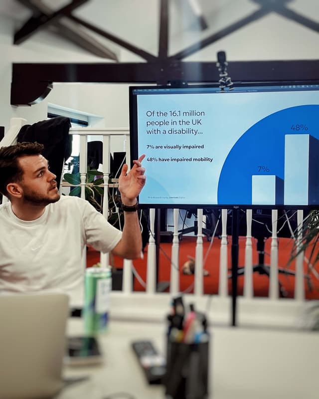 A man is talking and pointing at a screen with text on it saying 'Of the 16.1 million people in the UK with a disability... 7% are visually impaired, 48% have impaired mobility'