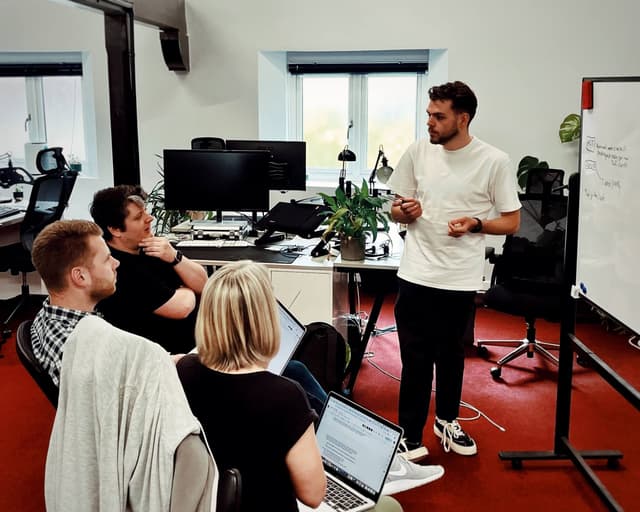 A man is standing up, casually dressed in a white t shirt and jeans and trainers. He's at a whiteboard and speaking to three colleagues sitting down to the left of him, with laptops open. He's talking about how accessibility is crucial to any website and explaining how an affordable accessibility testing tool would make a difference to businesses.