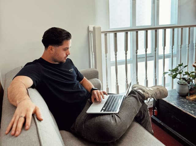 A man with dark hair and a t shirt is sitting on a sofa next to bannisters in front of a window. He has a laptop on his lap and am arm on the back of the sofa. He is looking at his laptop intently.