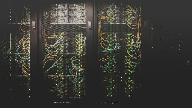 A close-up image of a server rack, featuring multiple rows of servers behind a mesh panel. The servers are connected with numerous cables, primarily in blue and orange, which weave in and out of the rack. Small green lights on the servers indicate activity, with some areas having brighter lights than others. The overall setting appears dark, highlighting the illuminated cables and lights, creating a high-tech atmosphere.