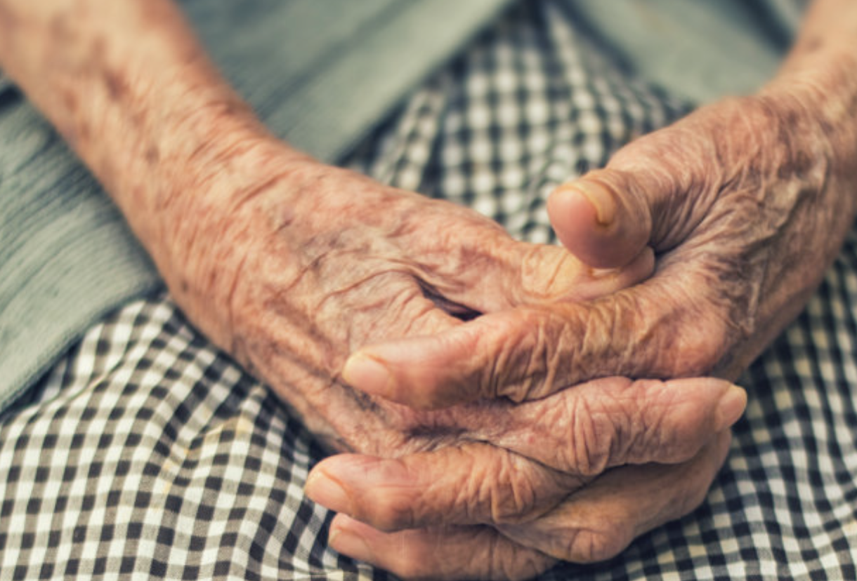 The image shows a close-up of two elderly hands resting on a checkered black and white fabric, likely a skirt or tablecloth. The skin is wrinkled and weathered, indicating age, with prominent veins and textures. The hands are clasped together, with the fingers interlaced and the thumb resting on top. The background is blurred, suggesting a soft focus on the hands and their details.