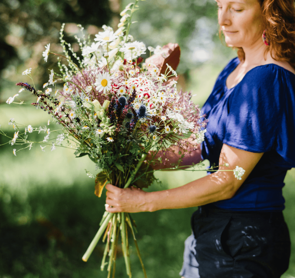 A woman in blue stands to the right, looking dreamily at a beautiful big bouquet she is holding of eco-friendly grown flowers and foliage